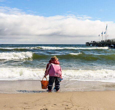 Kind spielt am Strand bei Wind und wetterfest, im Hintergrund Seebrücke