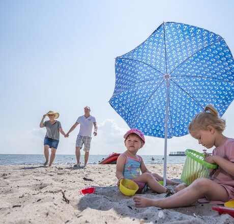 Kinder spielen mit Förmchen unter einem Sonnenschirm am Strand, im Hintergrund Eltern und das Meer