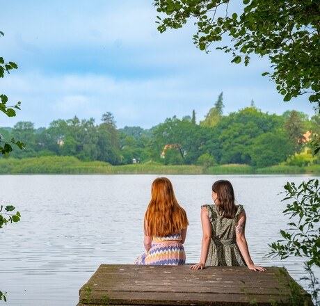 Zwei junge Frauen sitzen auf eine Steg am See und schauen auf die Natur der gegenüberliegenden Seite