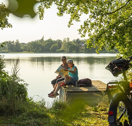 Ein Paar sitzend am See auf einem Steg, im Vordergrund ein Fahrrad
